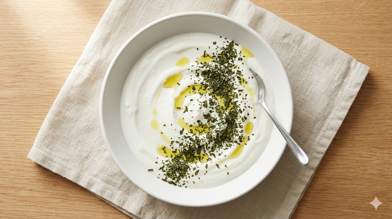 White ceramic bowl of yogurt sauce topped with dried mint and olive oil drizzle on a linen cloth
