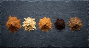 Five small piles of ground cinnamon, ginger, nutmeg, cloves and allspice arranged on a dark slate board before blending.